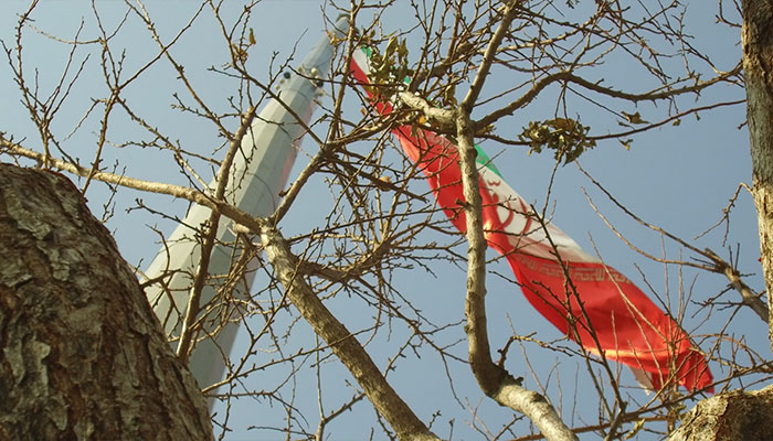 لانگ شات سینمایی از پرچم ایران از پشت درخت پاییزی در تهران