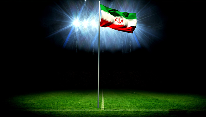 اهتزاز پرچم ملی ایران روی میله پرچم مقابل زمین فوتبال با نورافکن و فلاش